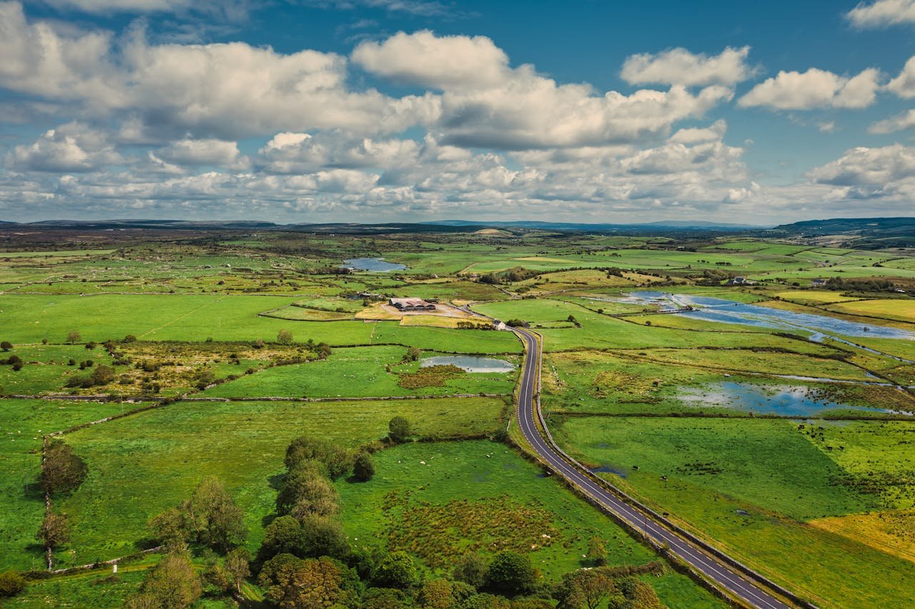 Aerial view of the vibrant green countryside and long road in County Clare, Ireland, under a partly cloudy sky.